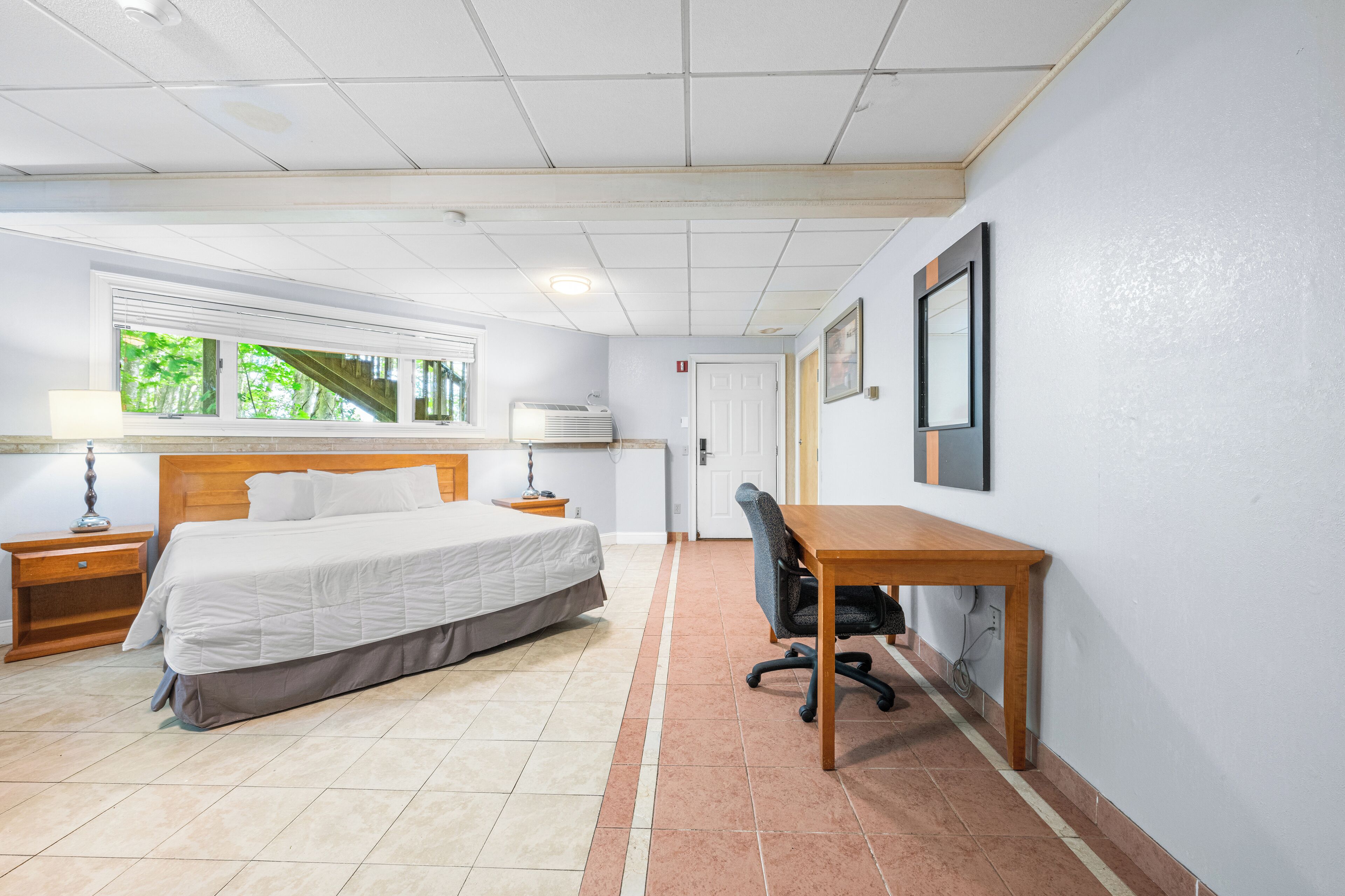 A hotel room with a bed, desk, chair, window, and a door.