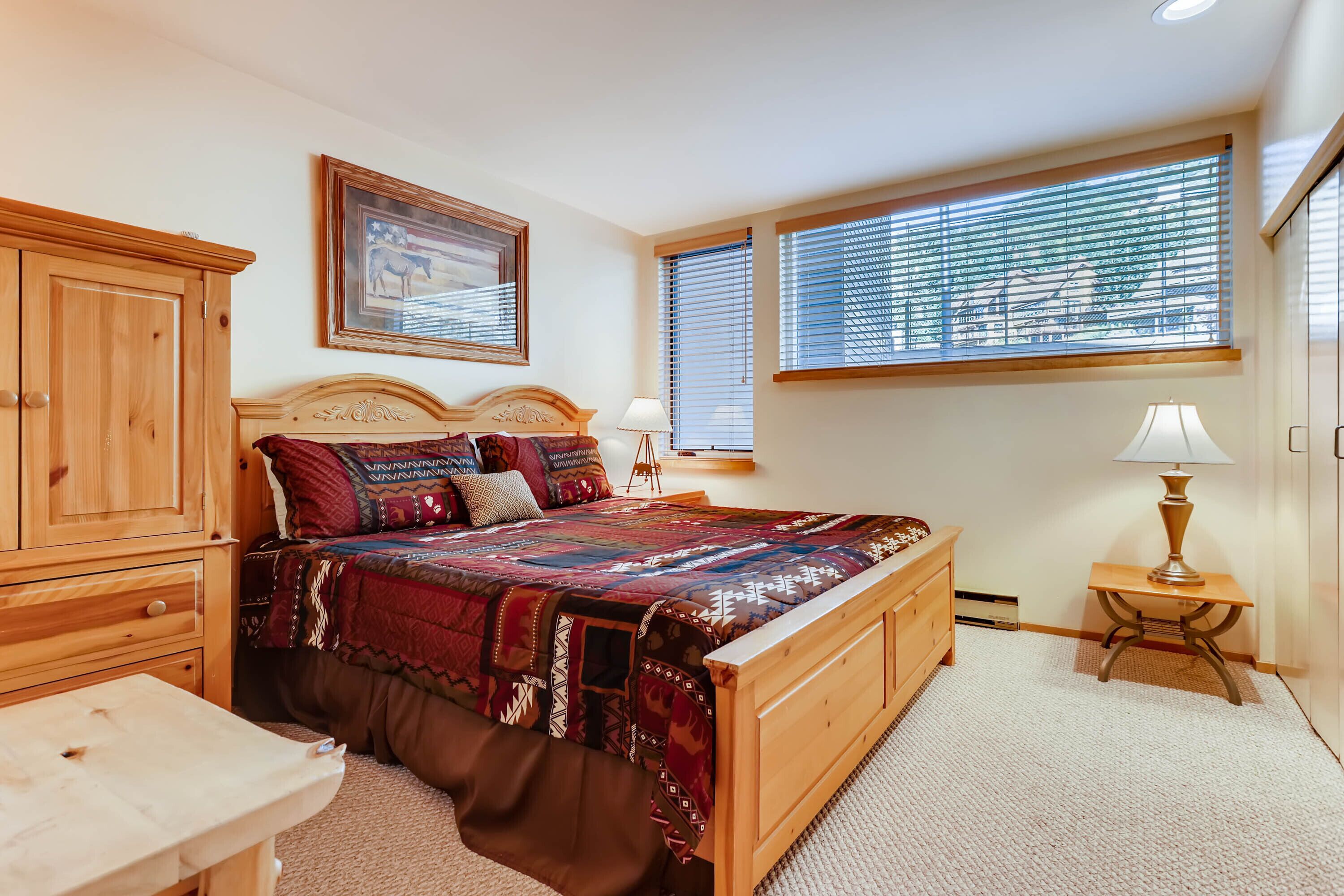 A bedroom with a wooden bed, a nightstand, a lamp, a wardrobe, and a window with blinds.