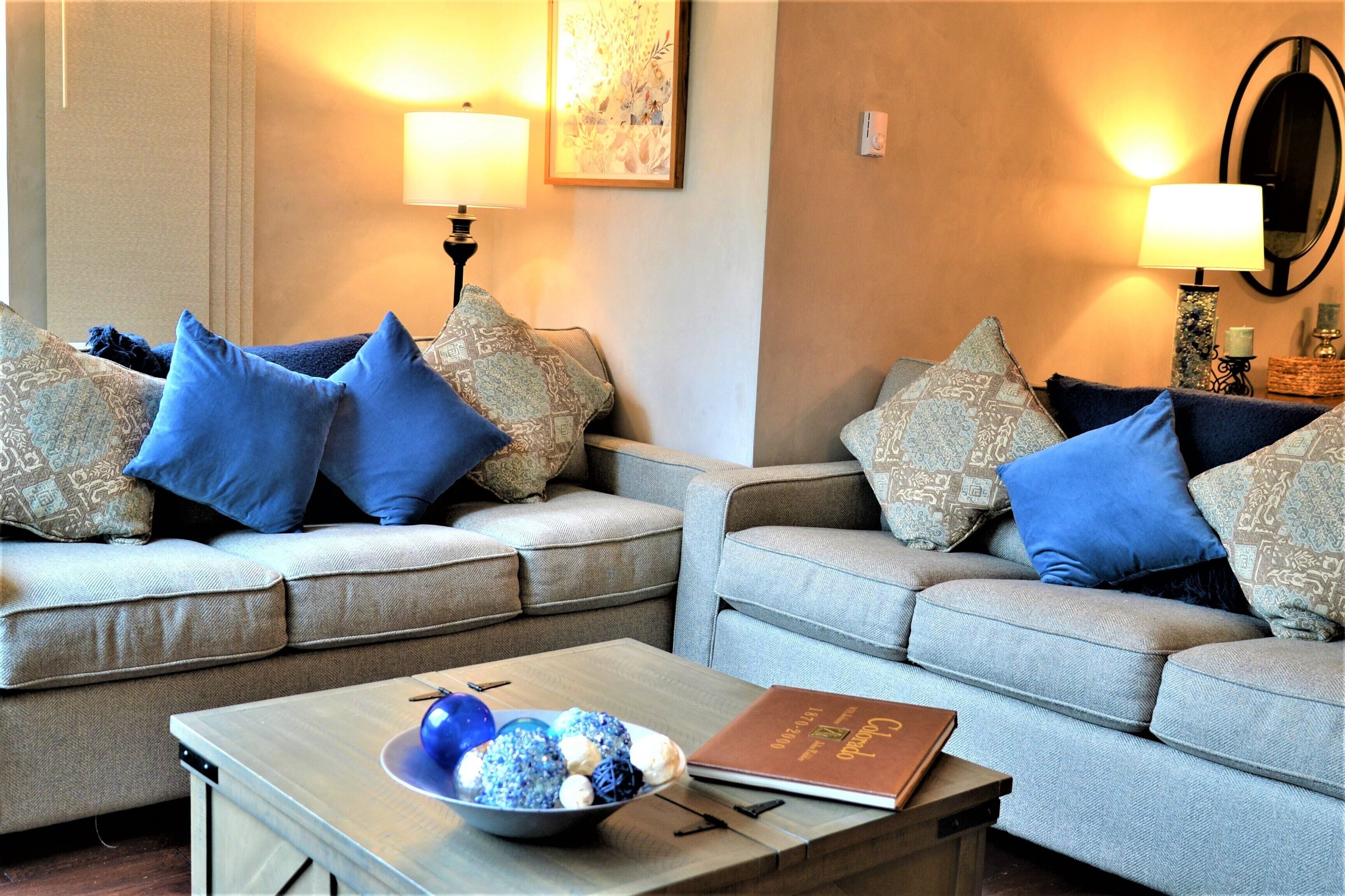 A living room with two grey sofas, each adorned with patterned cushions, a wooden coffee table with a bowl of decorative items, and a book.