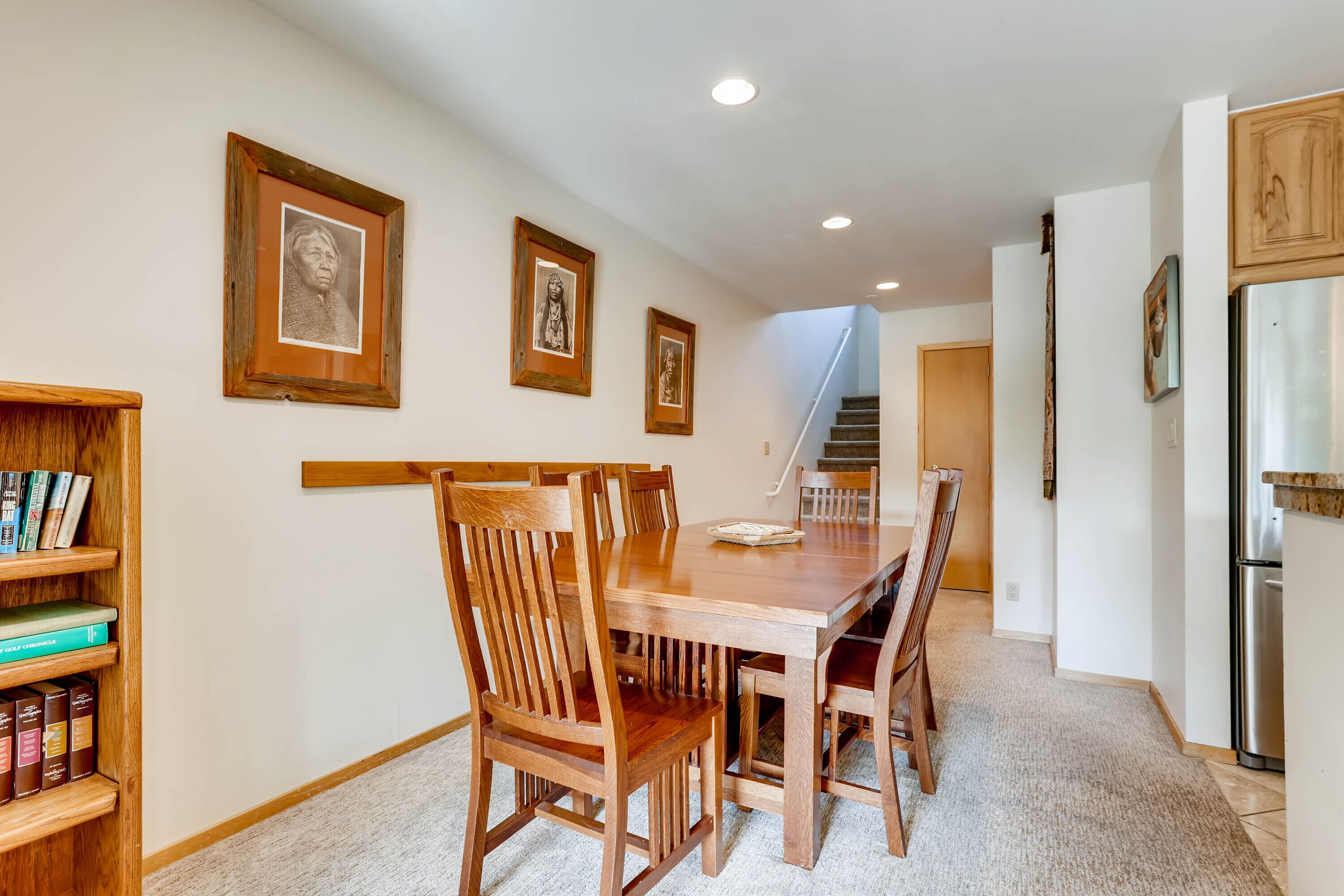 A dining area with a wooden table and chairs, framed pictures on the wall, and a staircase in the background.