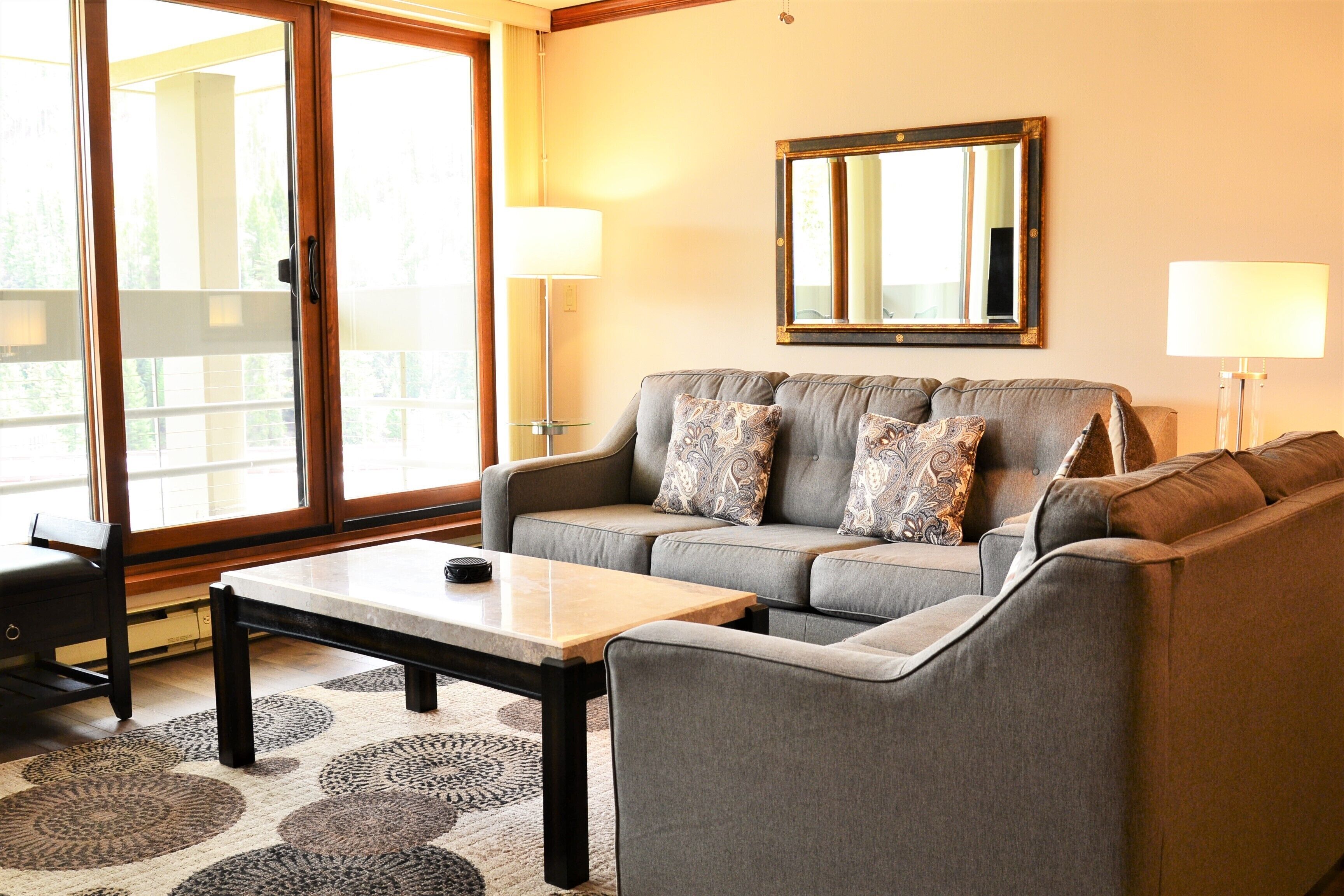 A living room with a grey sofa, two patterned throw pillows, a wooden coffee table, and a large mirror on the wall.