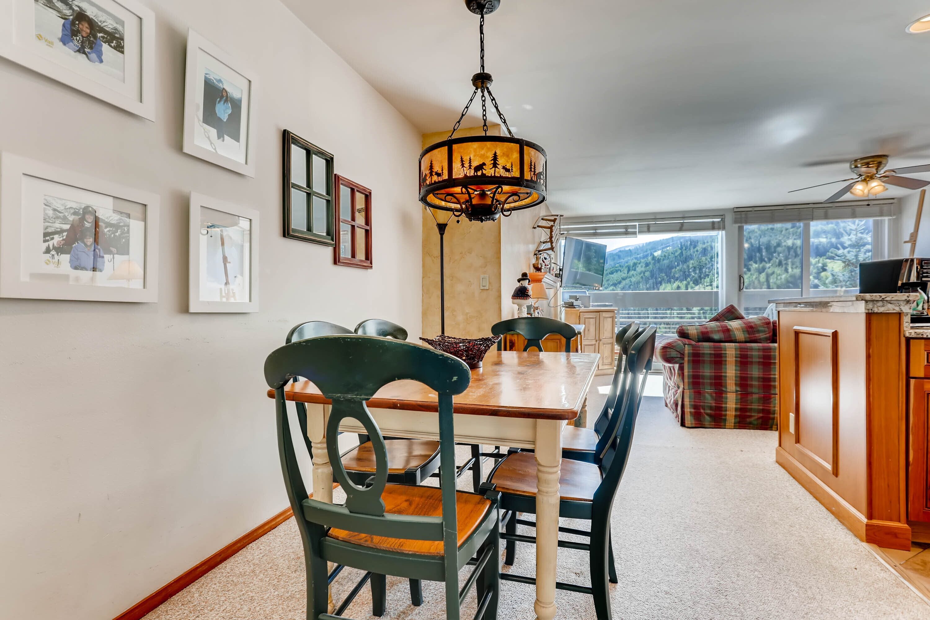 A dining area with a wooden table and chairs, a chandelier, framed pictures on the wall, and a view of the outdoors through large windows.