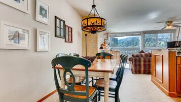 A dining area with a wooden table and chairs, a chandelier, framed pictures on the wall, and a view of the outdoors through large windows.