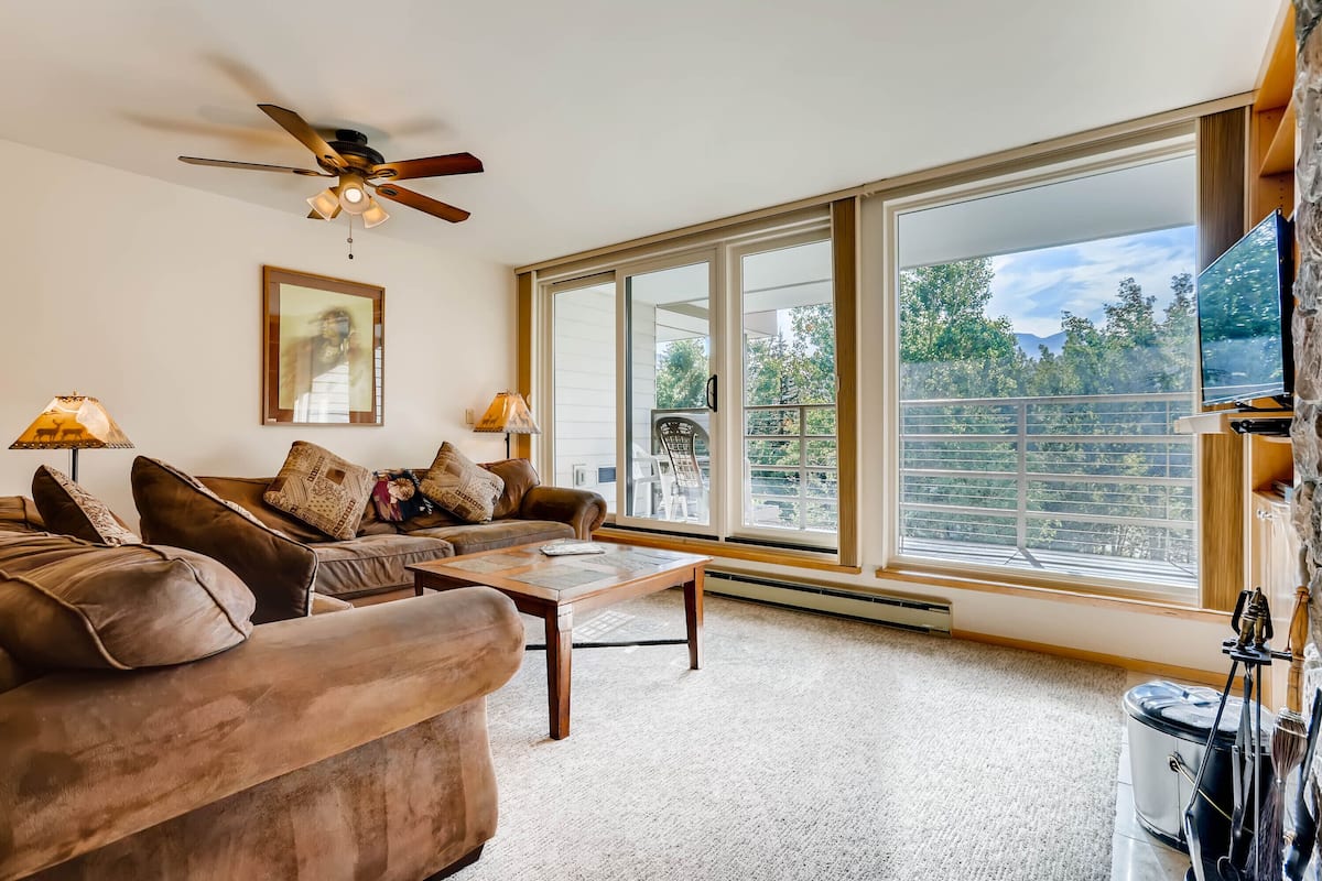 A spacious living room with a large sofa, a coffee table, and a ceiling fan. There is a large window offering a view of trees and a balcony.
