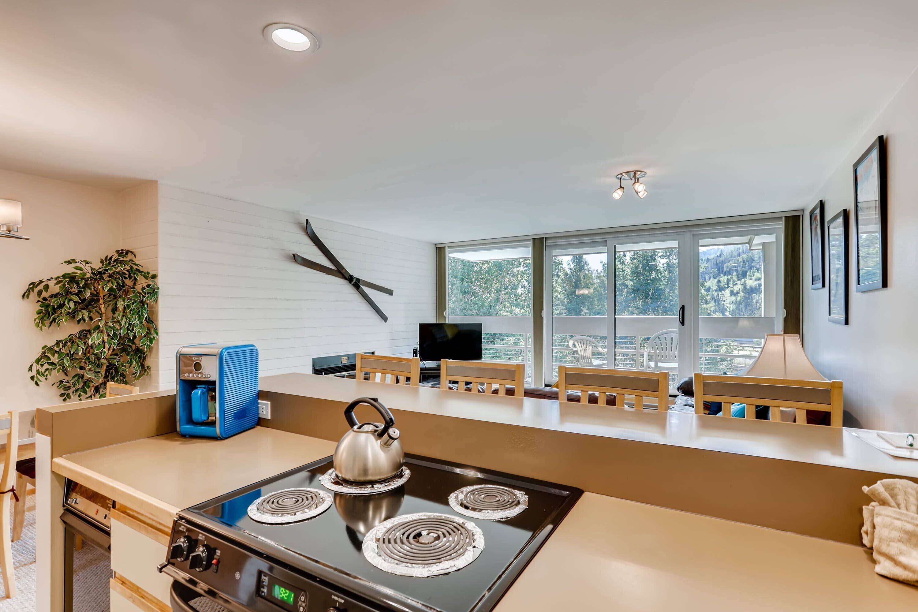 A modern kitchen with a stove, kettle, and a coffee machine. There are wooden chairs and a large window with a view of trees.