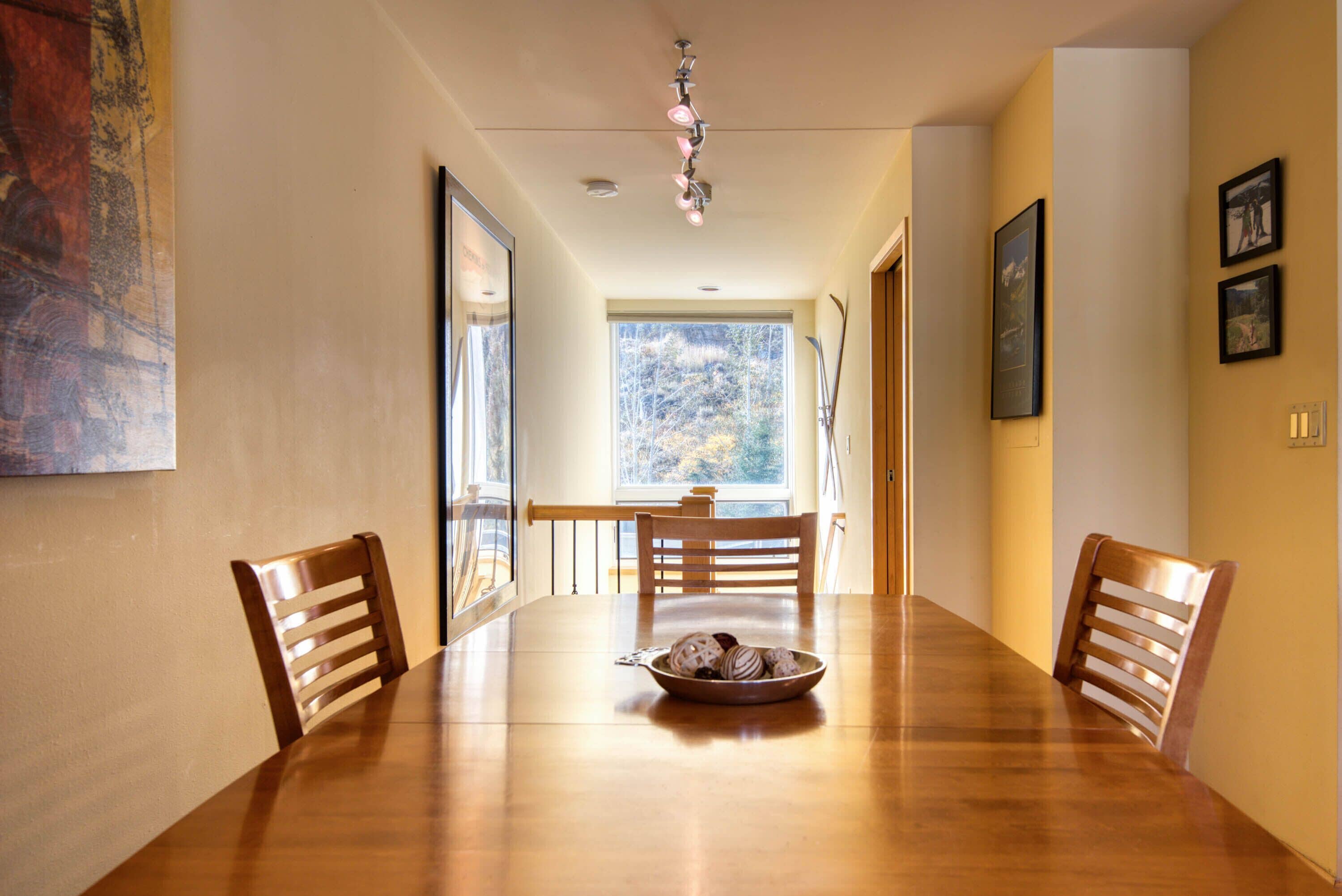 A dining room with a wooden table, four chairs, and a bowl of cookies. There is a window with a view of trees and a door leading to another room.