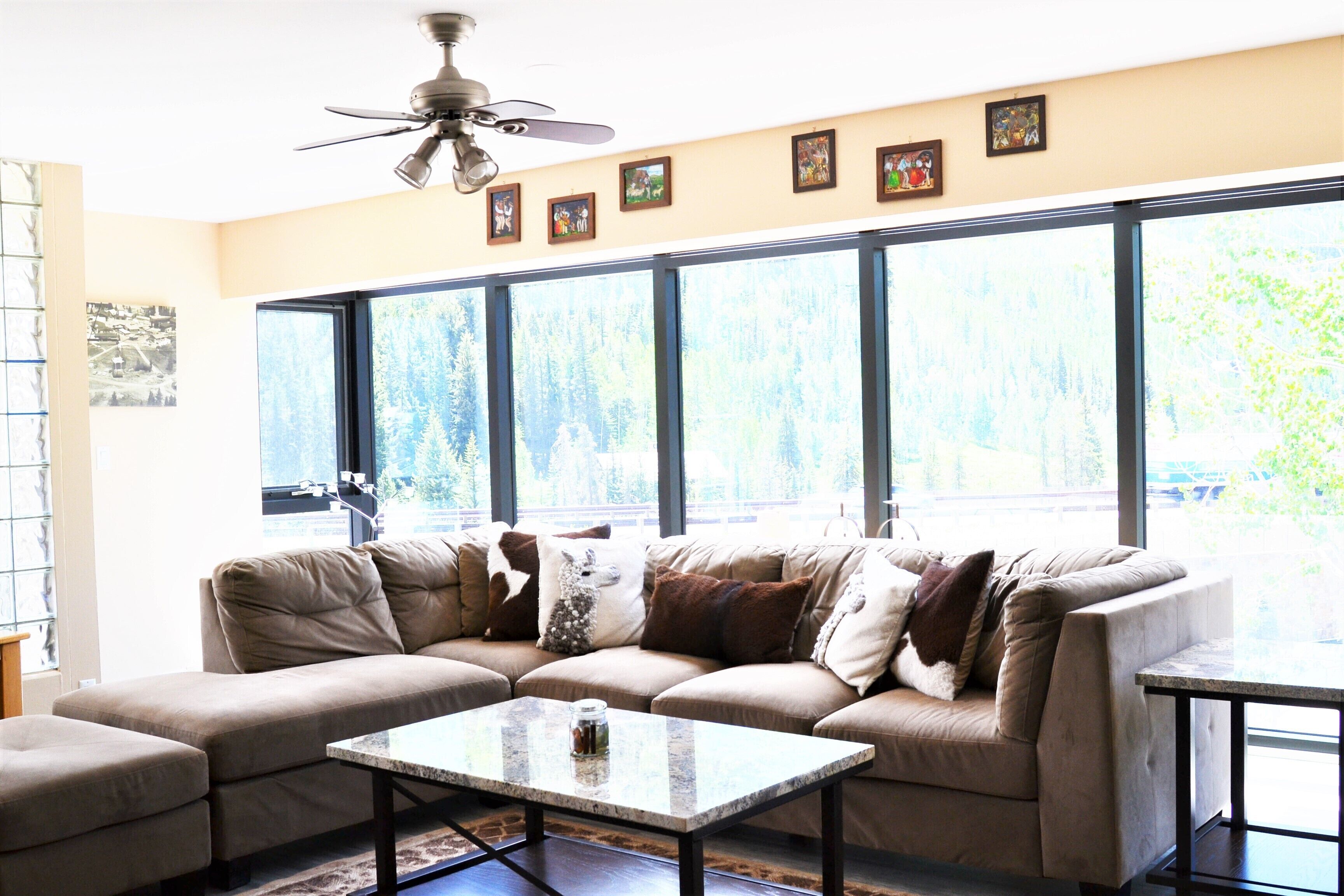 A living room with a sectional sofa, a coffee table, and large windows offering a view of trees.