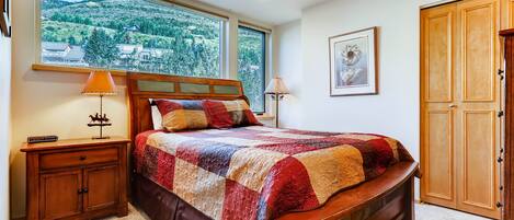 Un dormitorio con un ventanal grande, una cama con colcha, una mesita de noche, una lámpara, un ventilador de techo y un armario de madera.