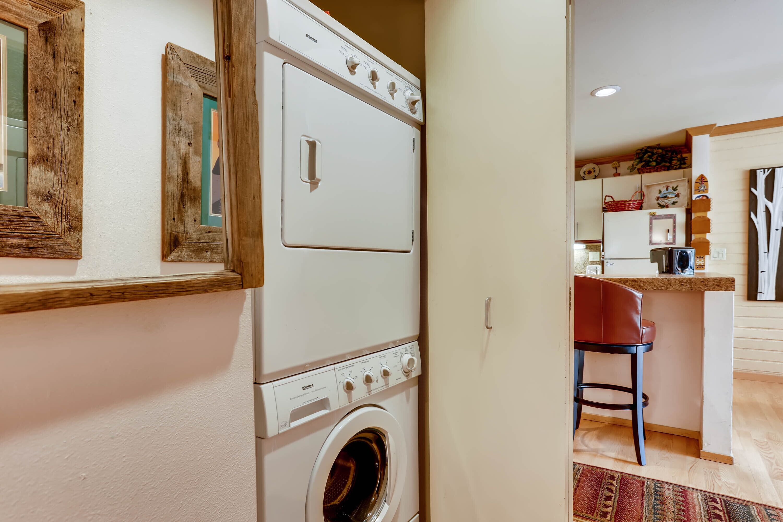 A laundry area with a front-loading washing machine and dryer, a wooden-framed mirror, and a kitchen area with a bar stool and countertop.