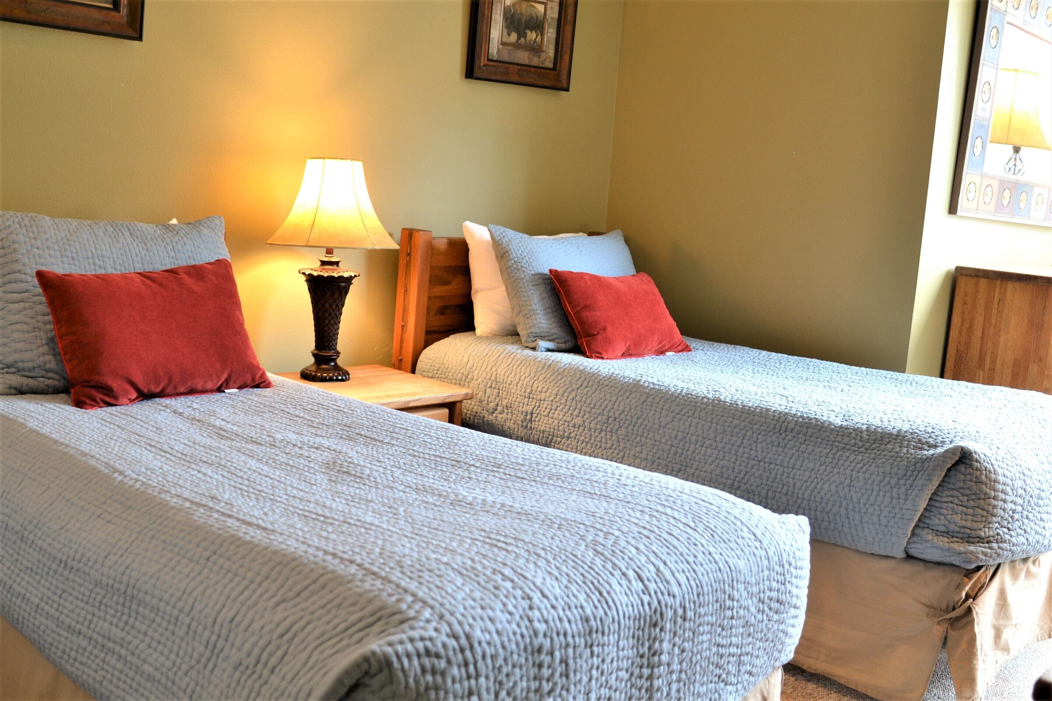 A hotel room with two single beds, each with a red pillow and a grey blanket. There is a nightstand with a lamp between the beds.