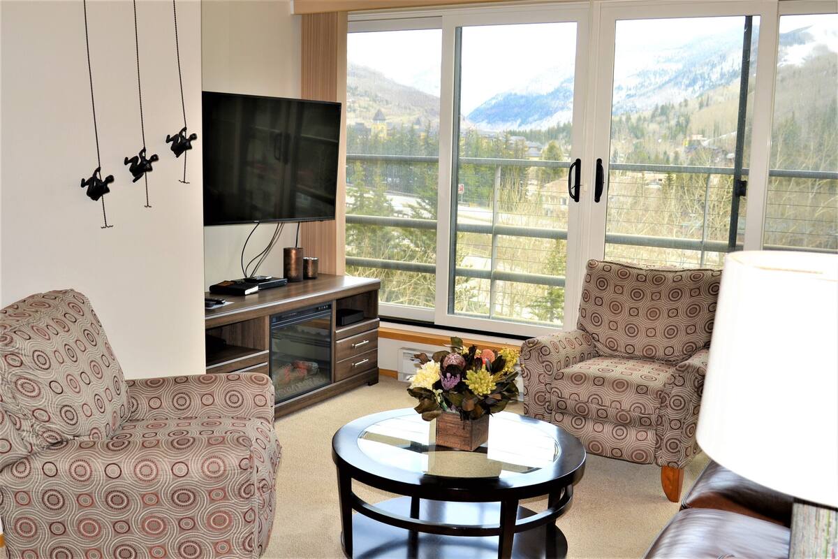 A living room with a TV, patterned chairs, a coffee table with flowers, and a view of mountains through large windows.
