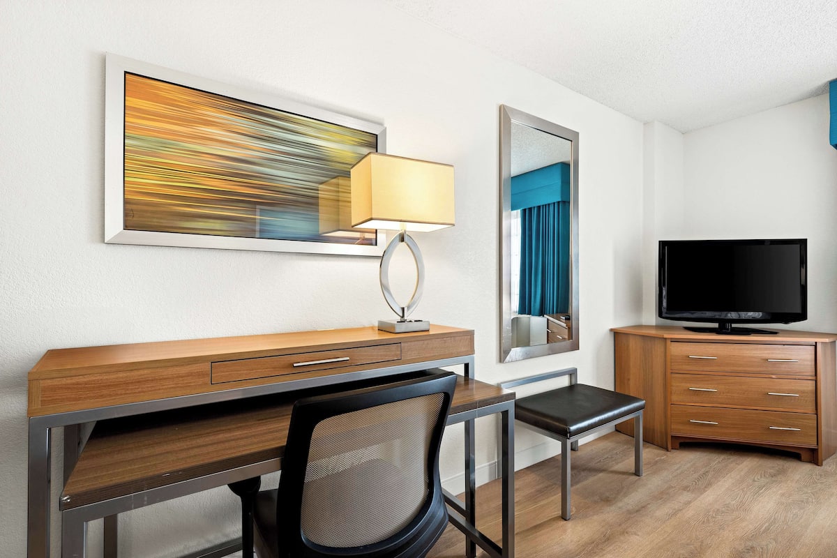A hotel room with a wooden desk, a chair, a television, a mirror, and a painting on the wall.