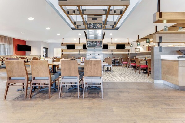 A spacious hotel dining area with multiple tables and chairs, a central staircase, and a modern kitchen area.