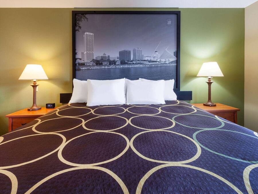A hotel room with a bed, two bedside lamps, a cityscape poster on the wall, and a nightstand with a clock.