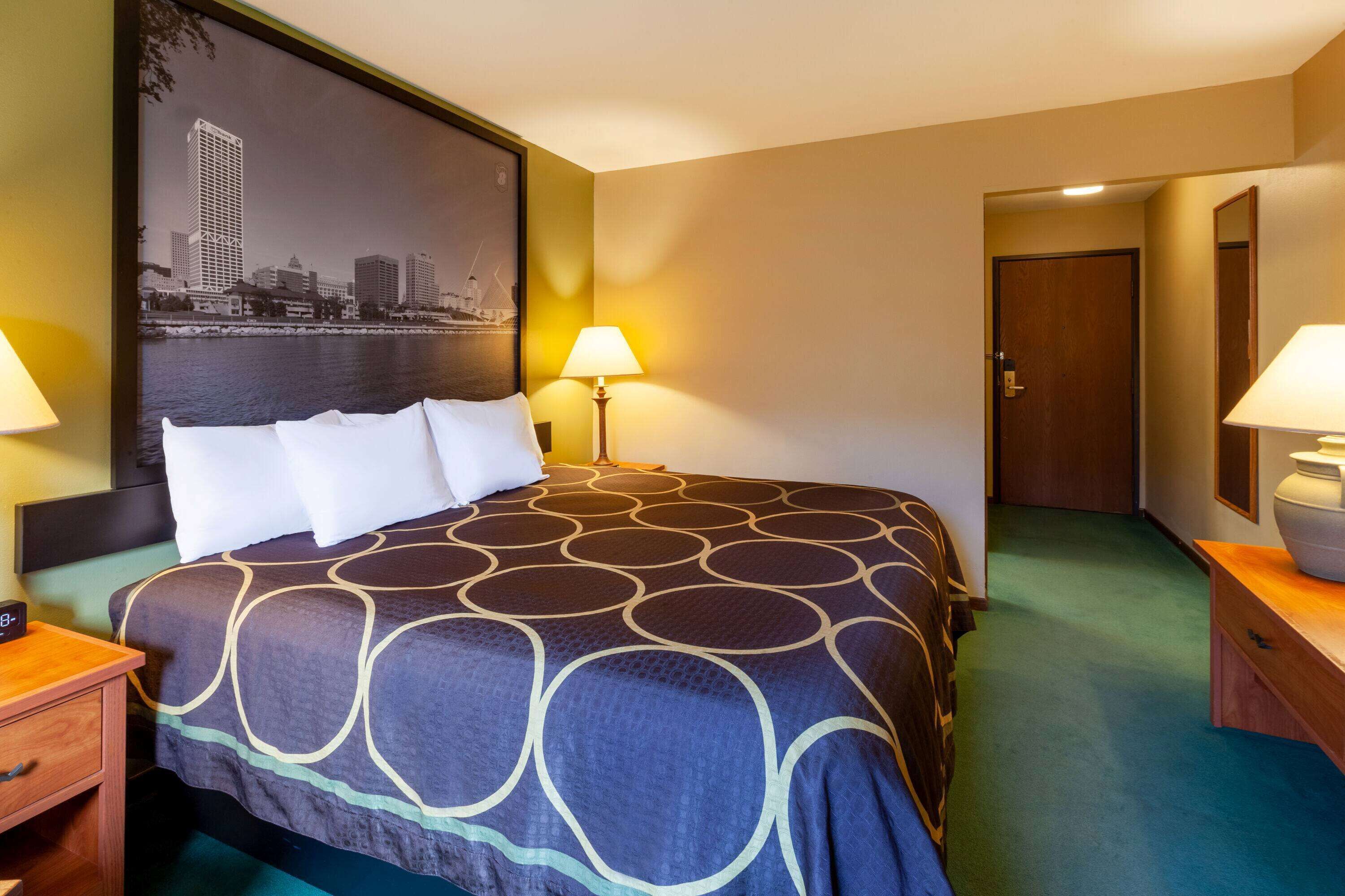 A hotel room with a large bed, two bedside lamps, a nightstand, a wooden dresser, and a door leading to another room.