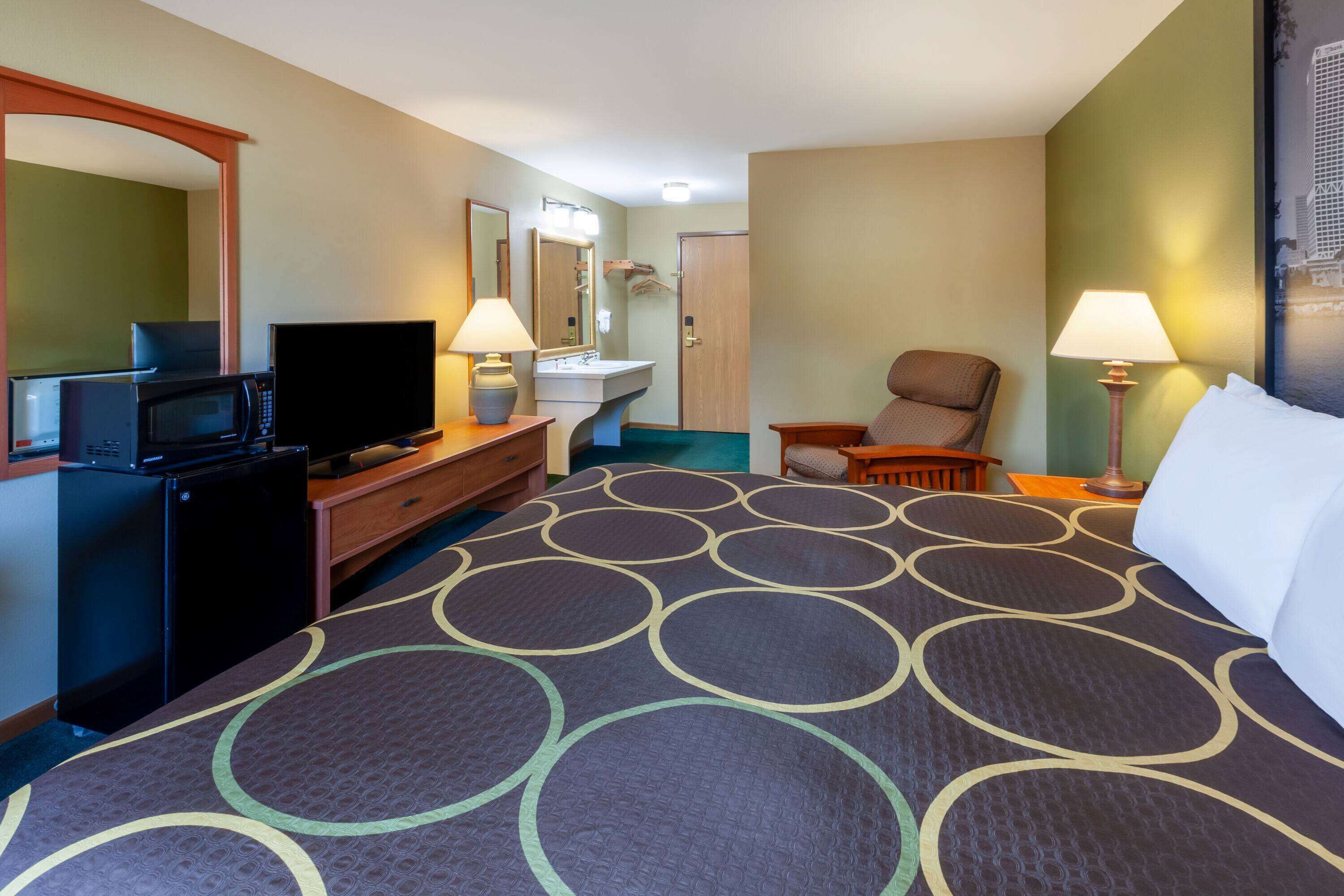 Habitación de hotel con cama, escritorio con televisor, microondas, silla y mesita de noche con lámparas.