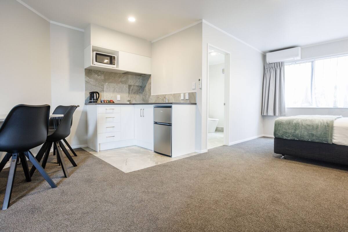 Cocina moderna con armarios blancos, un microondas y un refrigerador de acero inoxidable. Hay una zona de comedor con dos sillas negras y una pequeña mesa. La habitación tiene piso alfombrado y una cama con una manta verde.