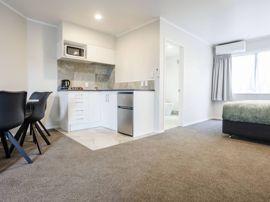 A modern kitchen with white cabinets, a microwave, and a stainless steel refrigerator. There is a dining area with two black chairs and a small table. The room has a carpeted floor and a bed with a green blanket.