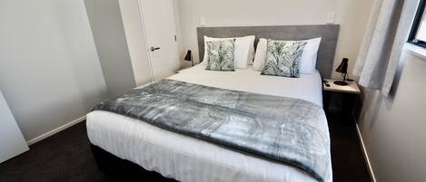 A neatly made bed with a grey and white patterned bedspread, white pillows, and a grey headboard. There is a bedside table with a lamp and a power outlet. The room has light grey walls and a dark carpet.