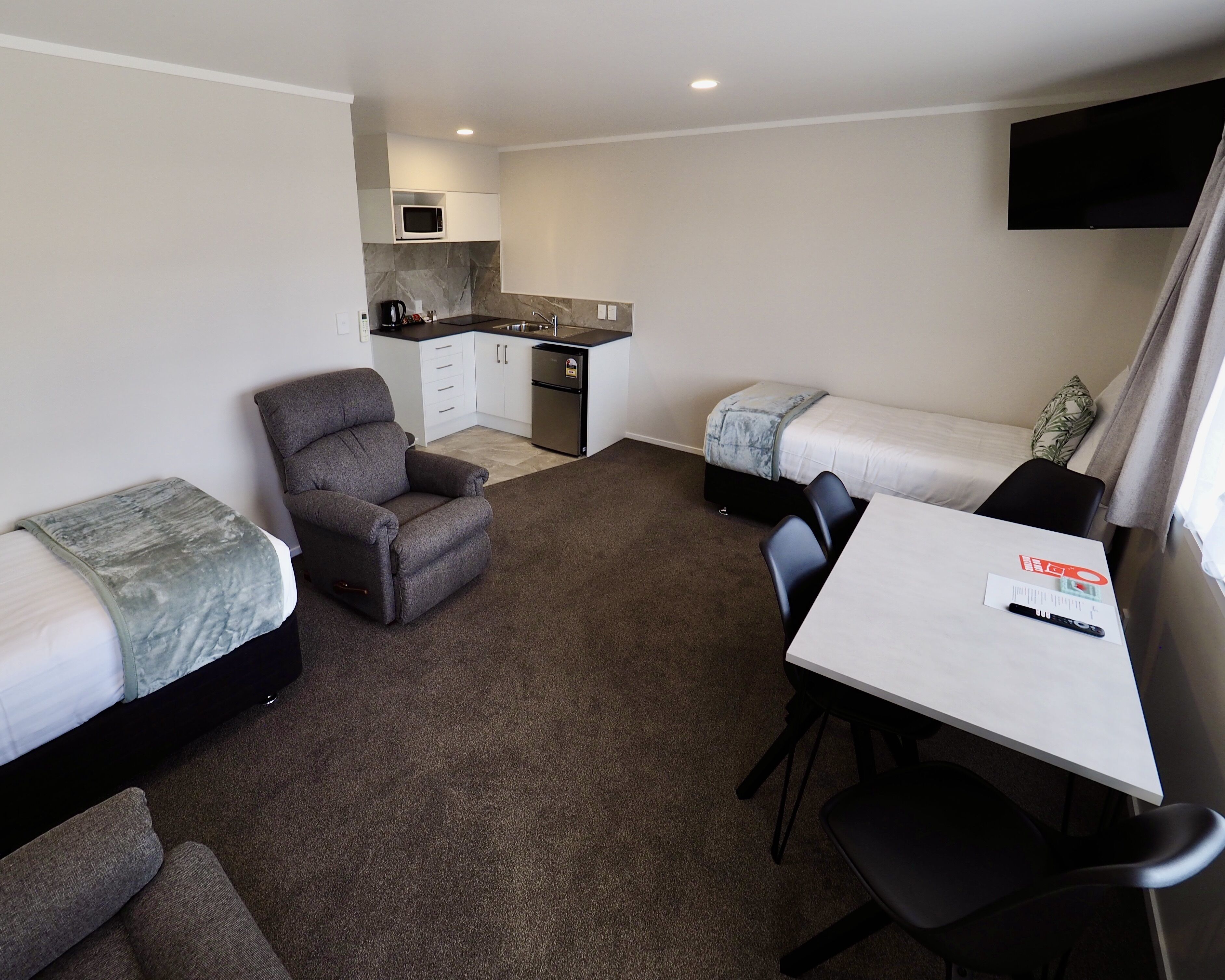 A compact room with two single beds, a small kitchenette, a sofa, a table with chairs, and a flat-screen TV mounted on the wall.