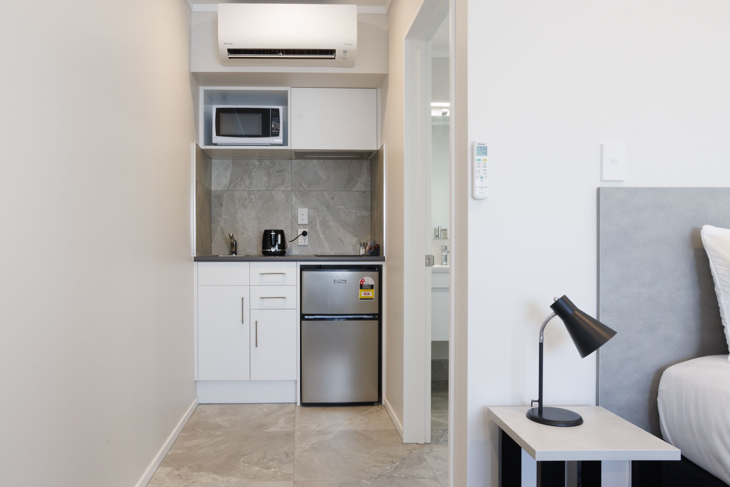 Cocina compacta con microondas, refrigerador y cafetera. Una mesita de noche con lámpara junto a la cama.