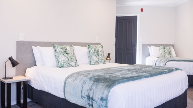 A hotel room with two beds, each with a grey headboard, white bedding, and green leaf-patterned pillows. There is a bedside table with a lamp on the left side. - Bk's Pohutukawa Lodge (Whangarei)