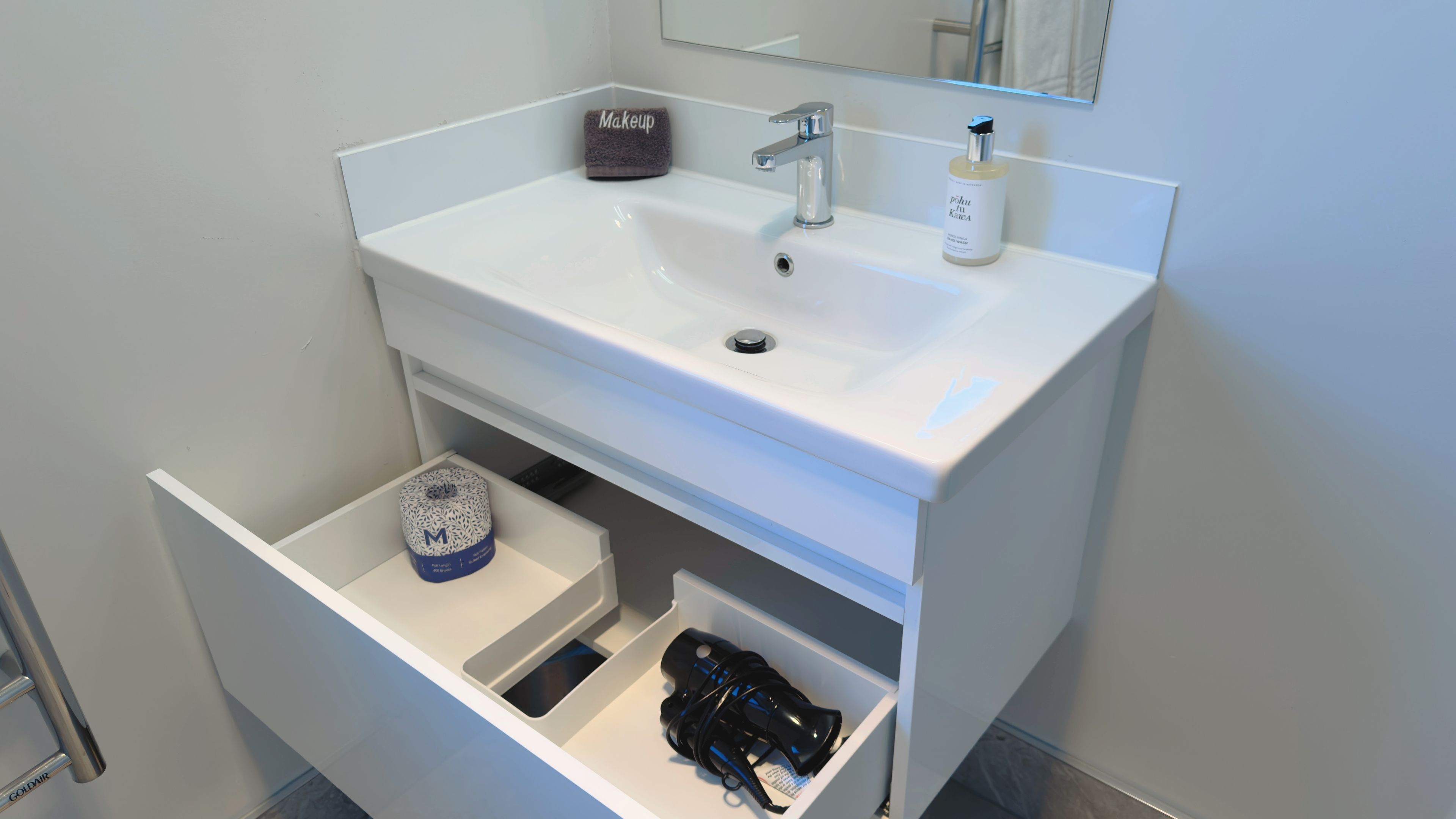 Une salle de bain avec un lavabo blanc, une serviette pour se maquiller, un flacon de savon liquide et un sèche-cheveux rangé dans un tiroir.