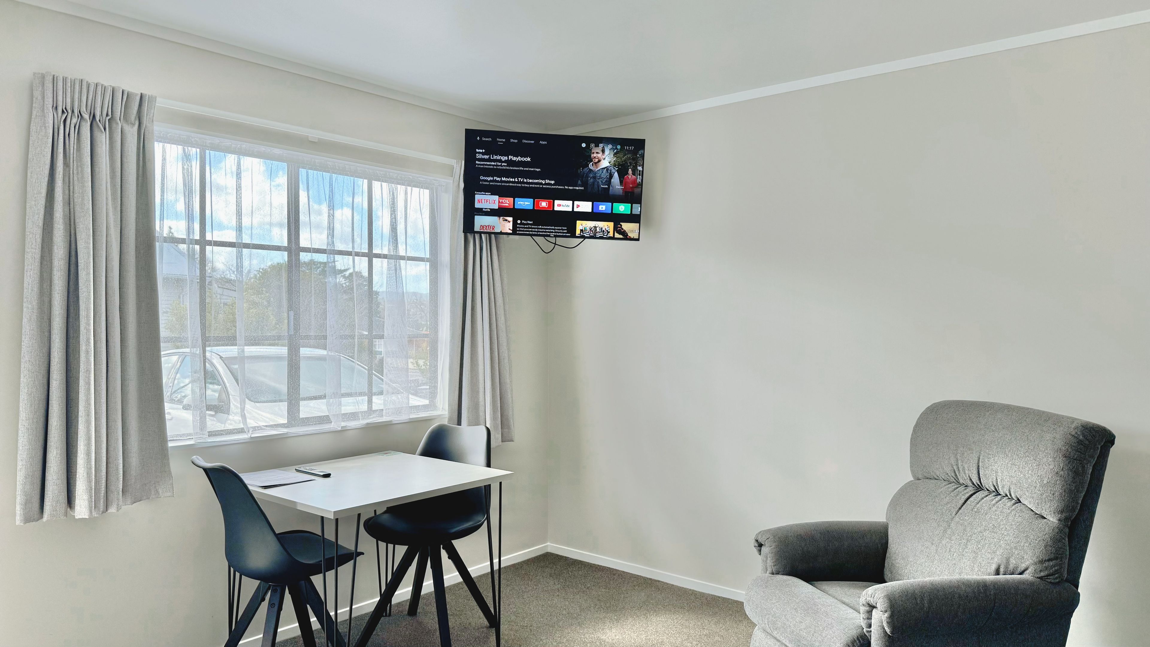 A room with a TV mounted on the wall, a table with two chairs, a grey sofa, and a window with curtains.
