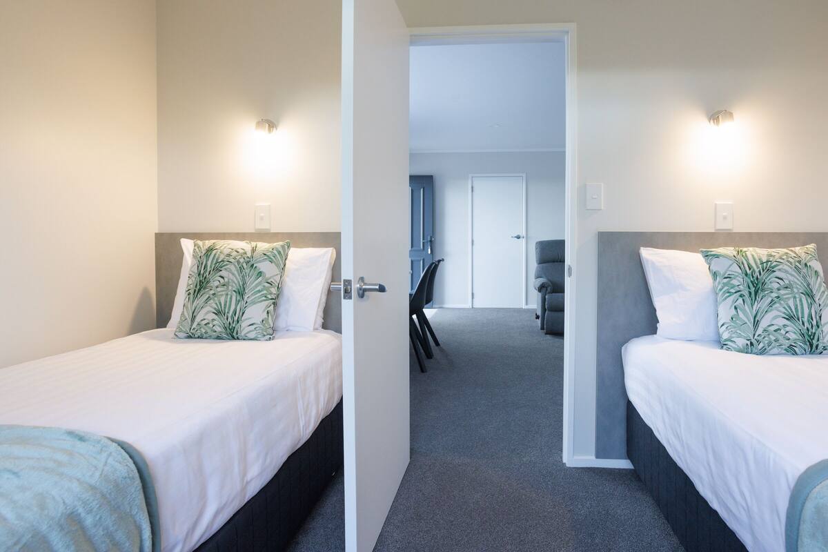 Habitación de hotel con dos camas, cada una con una almohada y una manta. Se ve una puerta que da a otra habitación.