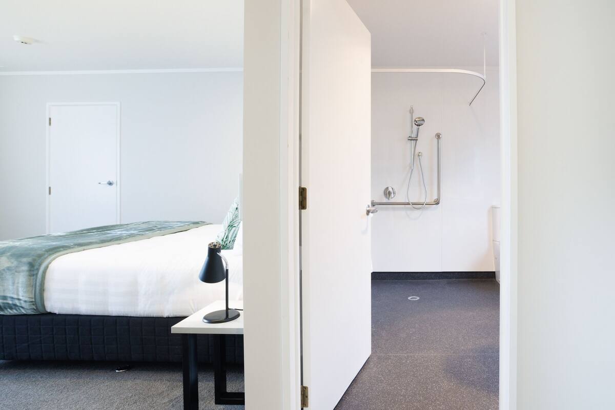 Una habitación de hotel con cama, escritorio con lámpara y zona de ducha.