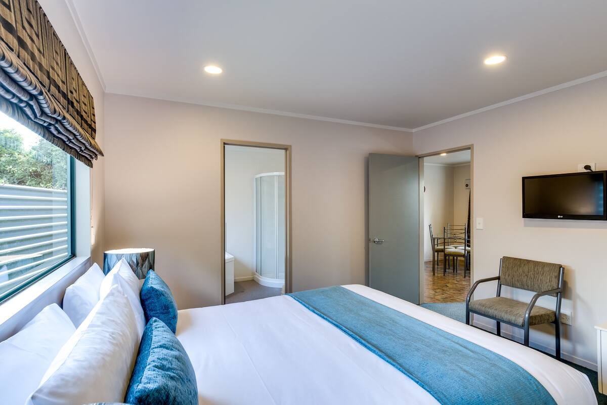 A hotel room with a large bed, a chair, a TV, and a window with blinds.