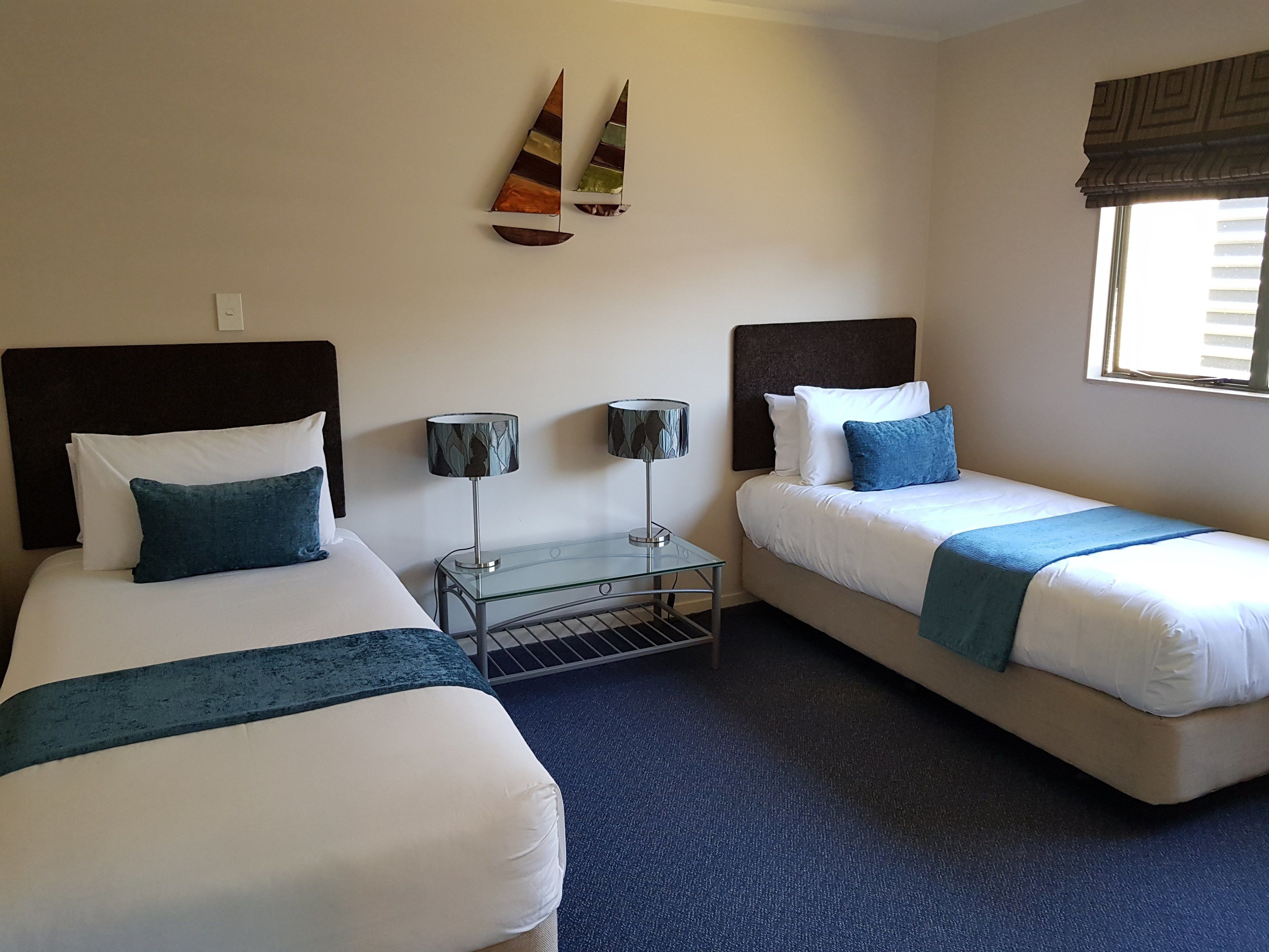 A hotel room with two beds, a small table, two lamps, and a window with blinds.