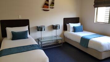 A hotel room with two beds, a small table, two lamps, and a window with blinds.