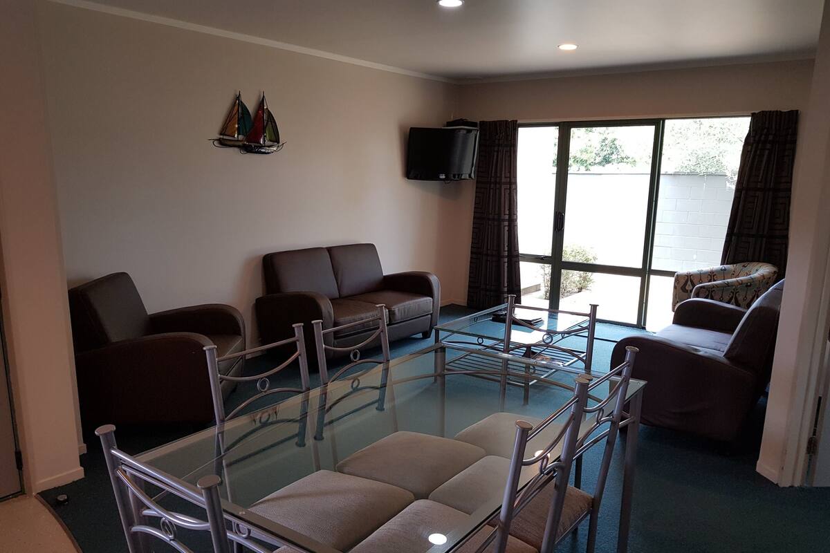 A room with a glass table surrounded by chairs, a sofa, and a television on the wall.