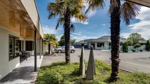 A modern building with a covered outdoor seating area, palm trees, and a parking lot. - Portmans Motor Lodge (Hastings)