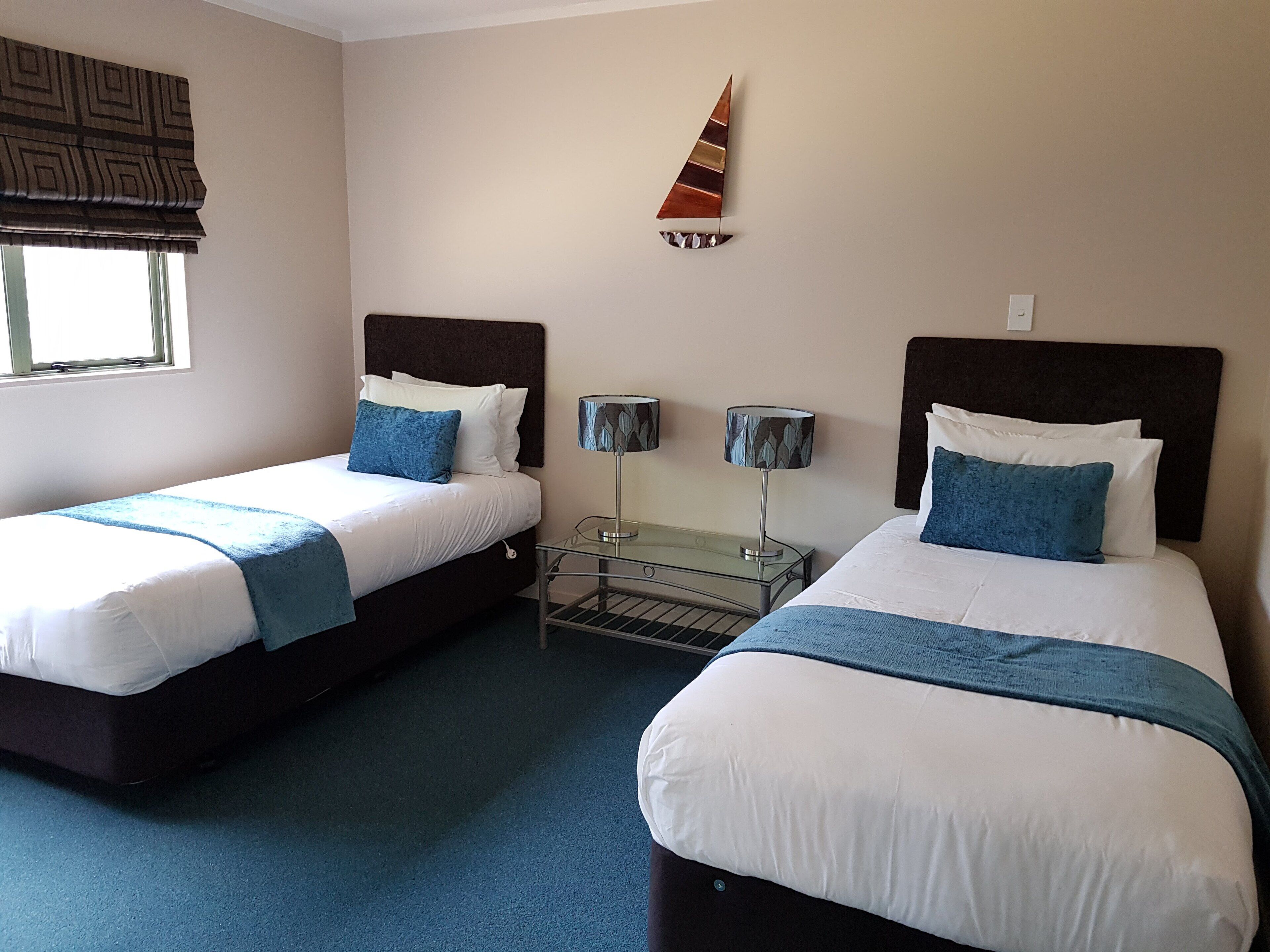 A hotel room with two beds, a window with a patterned blind, a wall-mounted decorative sail, and a small table with two lamps.