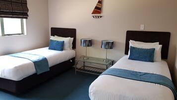 A hotel room with two beds, a window with a patterned blind, a wall-mounted decorative sail, and a small table with two lamps.