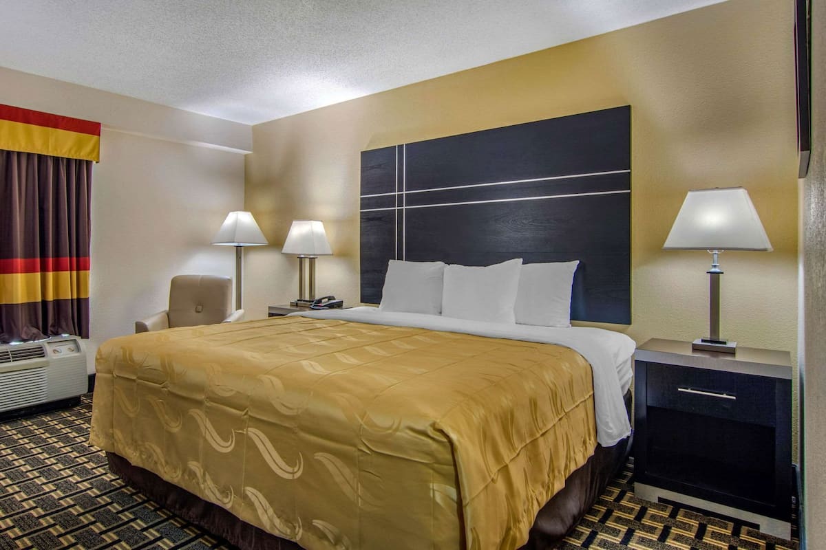 A hotel room with a large bed, two bedside lamps, a nightstand, an air conditioning unit, and a window with curtains.