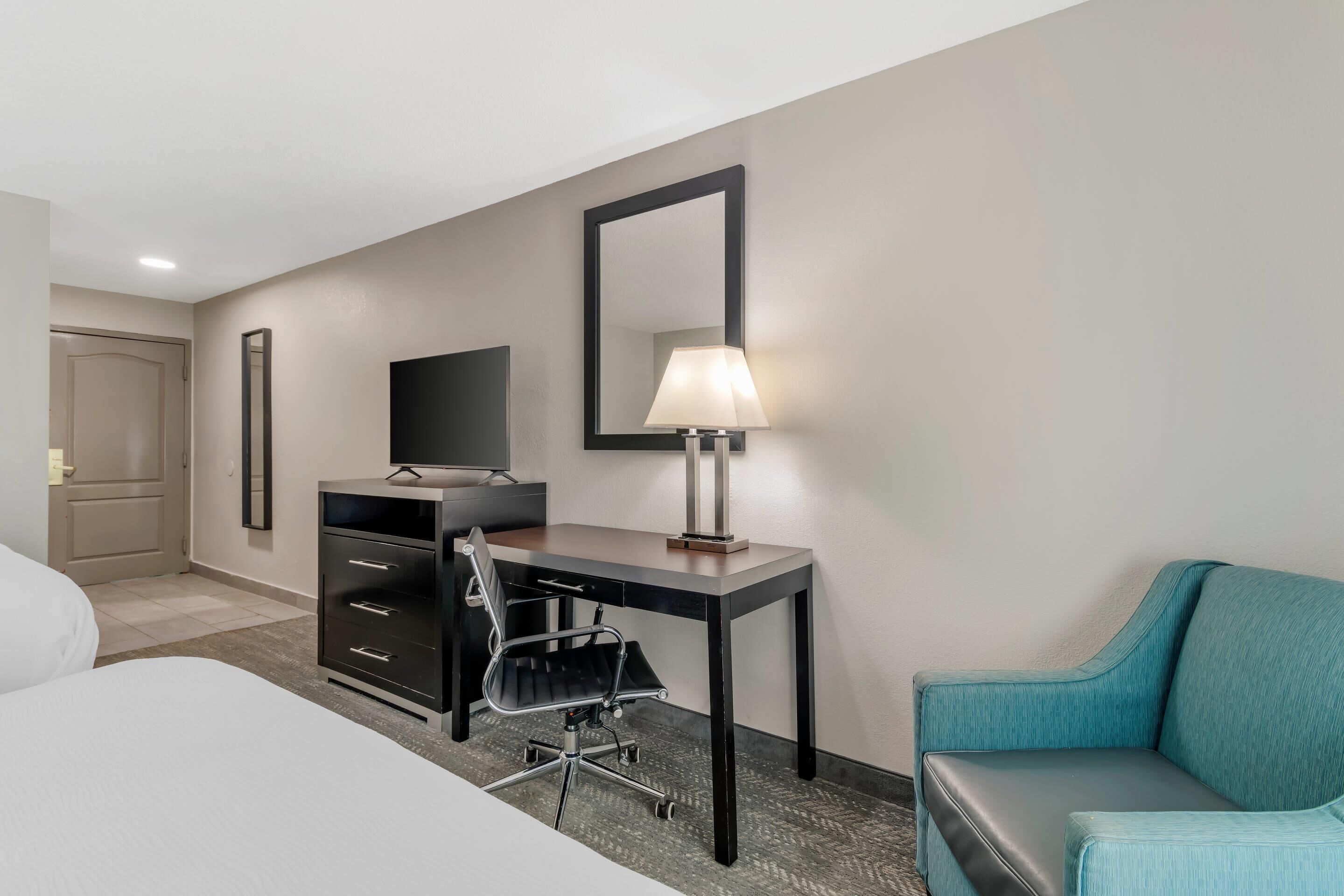 A hotel room with a bed, a desk with a chair, a television, a lamp, and a large mirror.