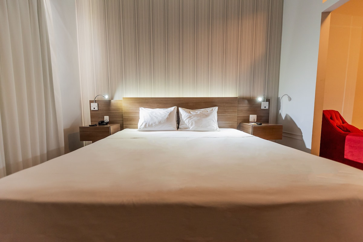 Una habitación de hotel con una cama grande, cabecera de madera, dos mesitas de noche con lámparas y una silla roja.