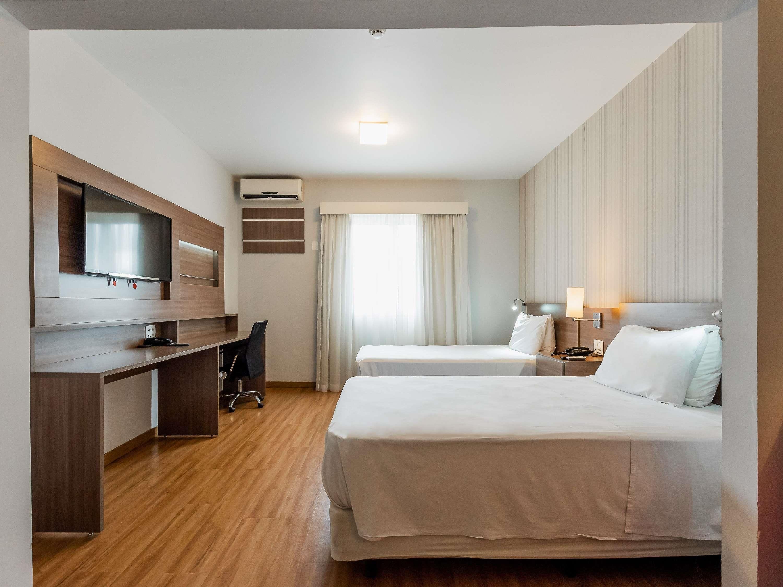 A hotel room with a wooden desk, a flat-screen TV, two beds, a desk lamp, and a window with curtains.