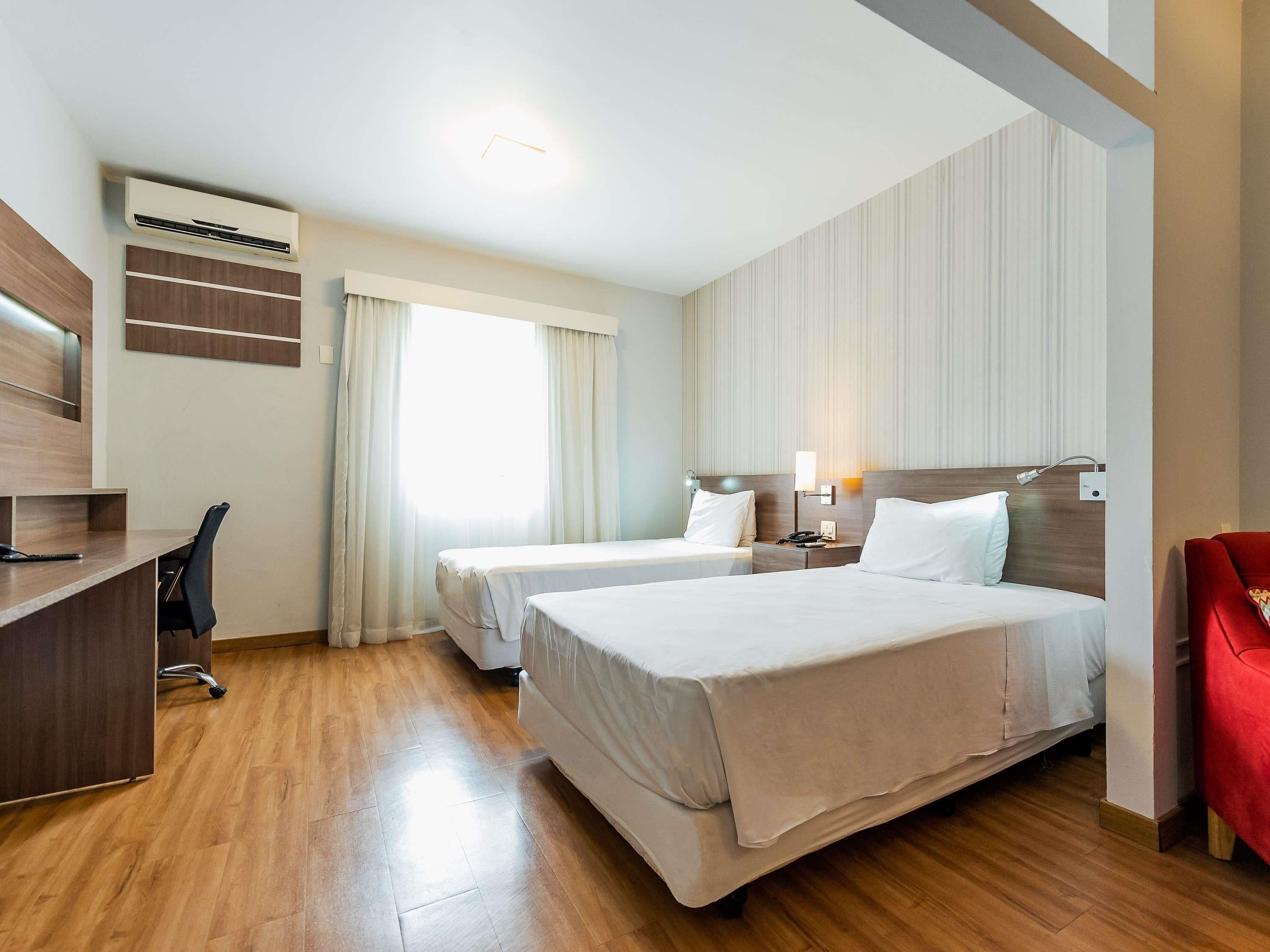 A hotel room with two beds, a desk, a chair, a lamp, and a window with curtains.