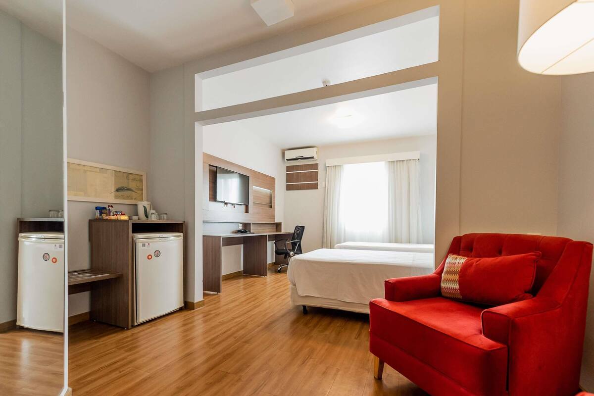 A hotel room with a bed, a red armchair, a desk with a computer, a kitchenette, and a TV.
