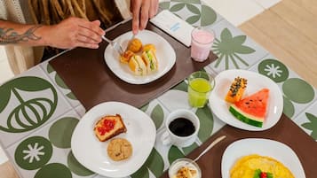 Mesa de café da manhã com pratos de comida, um copo de suco e uma xícara de café, e uma pessoa comendo.