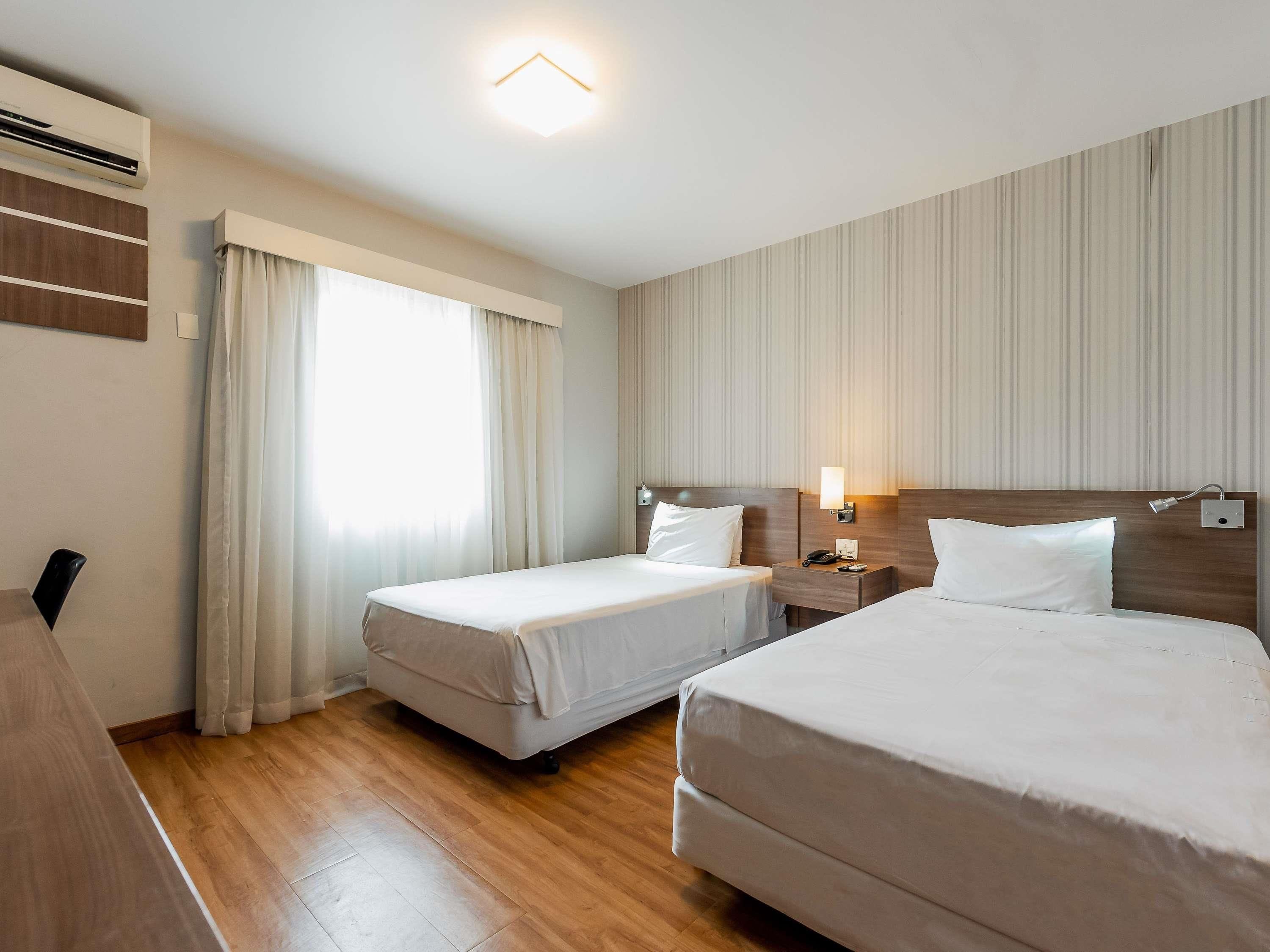 Habitació d'hotel amb dos llits, una taula de treball, una tauleta de nit, una làmpada i una finestra amb cortines.