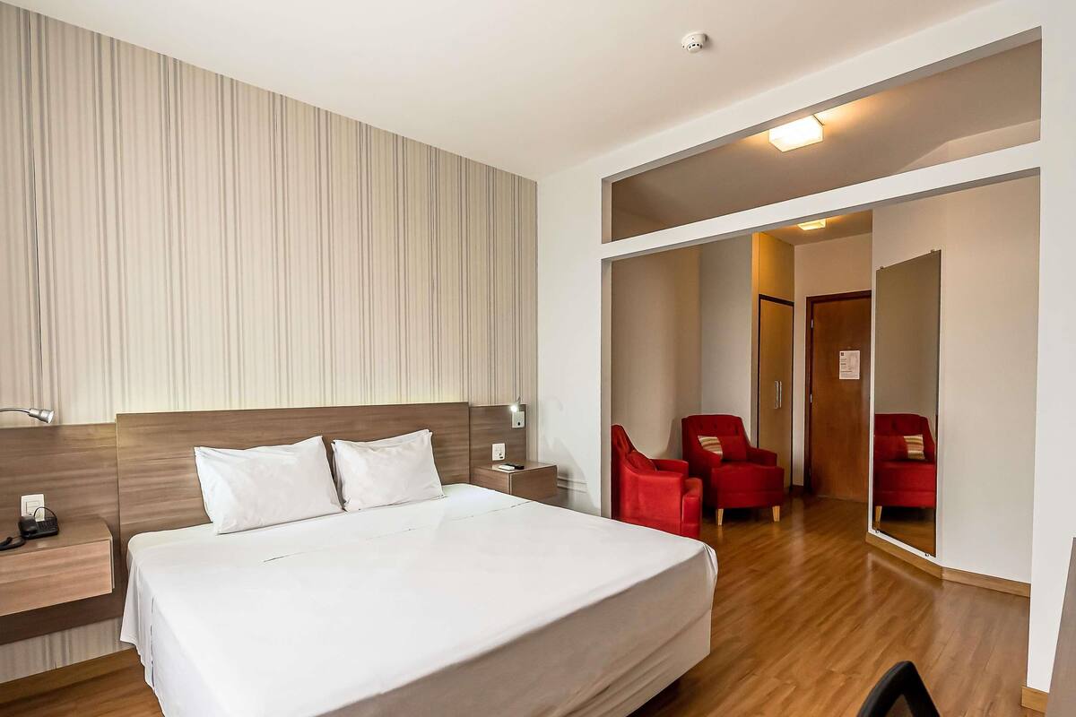 A hotel room with a large bed, wooden headboard, bedside tables with lamps, a red armchair, and a mirror reflecting the room.