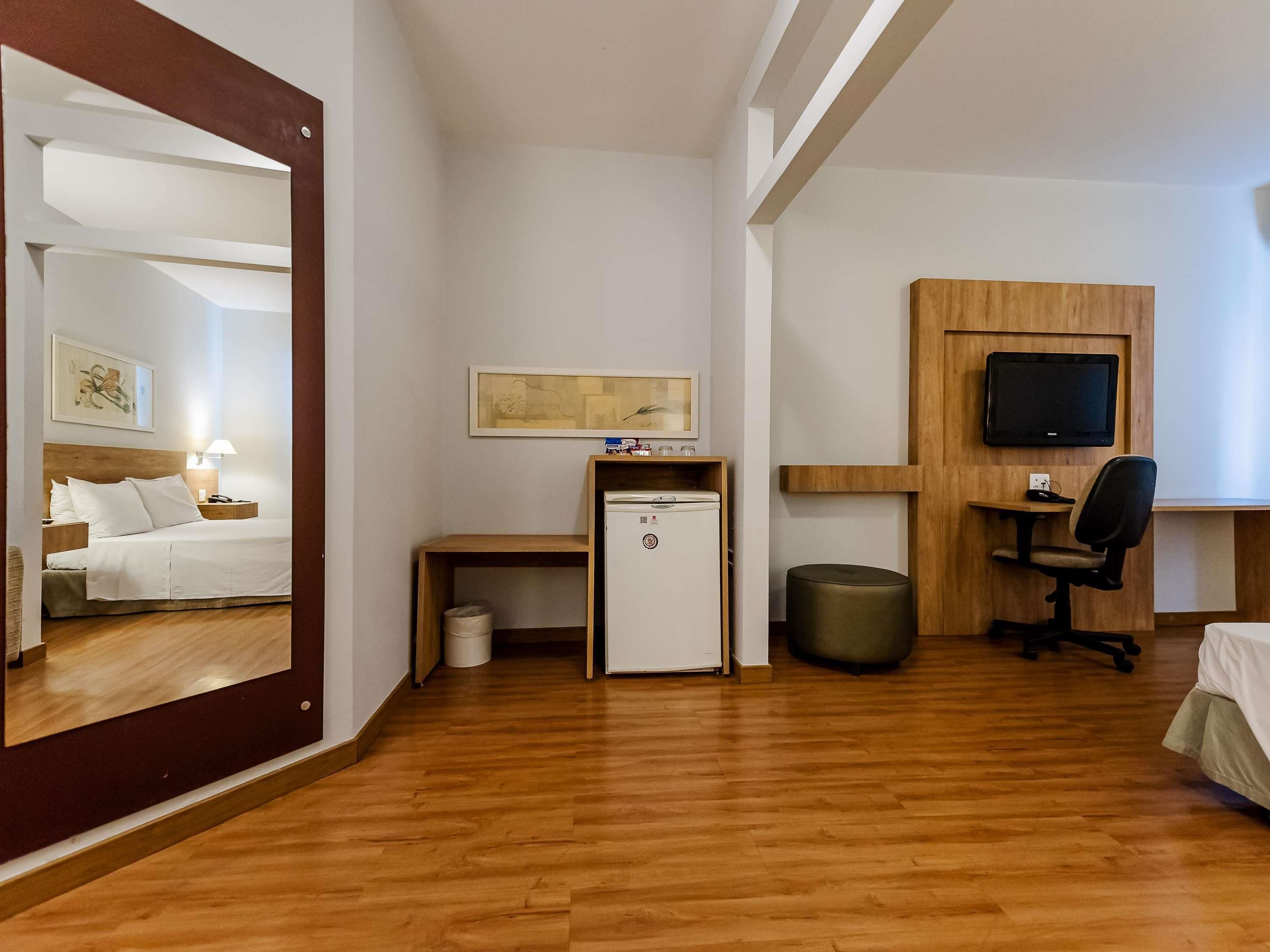 Habitación de hotel con cama, escritorio, refrigerador pequeño, cesto de basura, televisor y espejo.