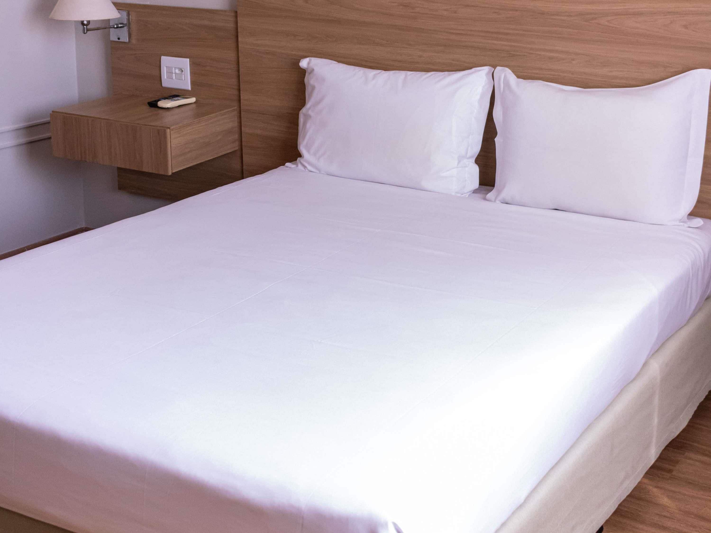 Una cama bien hecha con sábanas blancas, cabecera de madera, mesita de noche con lámpara y toma de corriente.