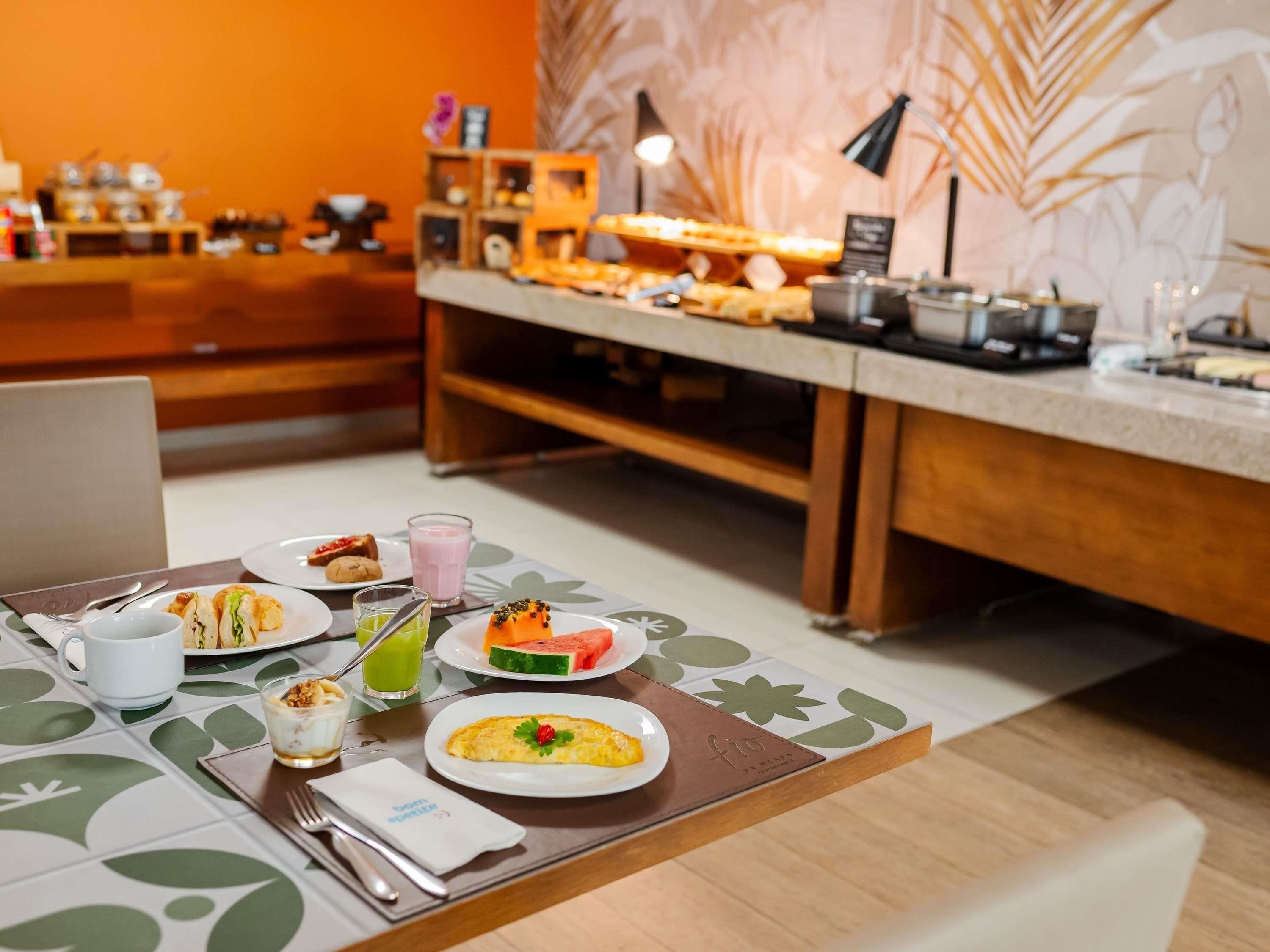 A breakfast buffet with various dishes and drinks, a laptop on the table, and a wooden counter with kitchen appliances in the background.