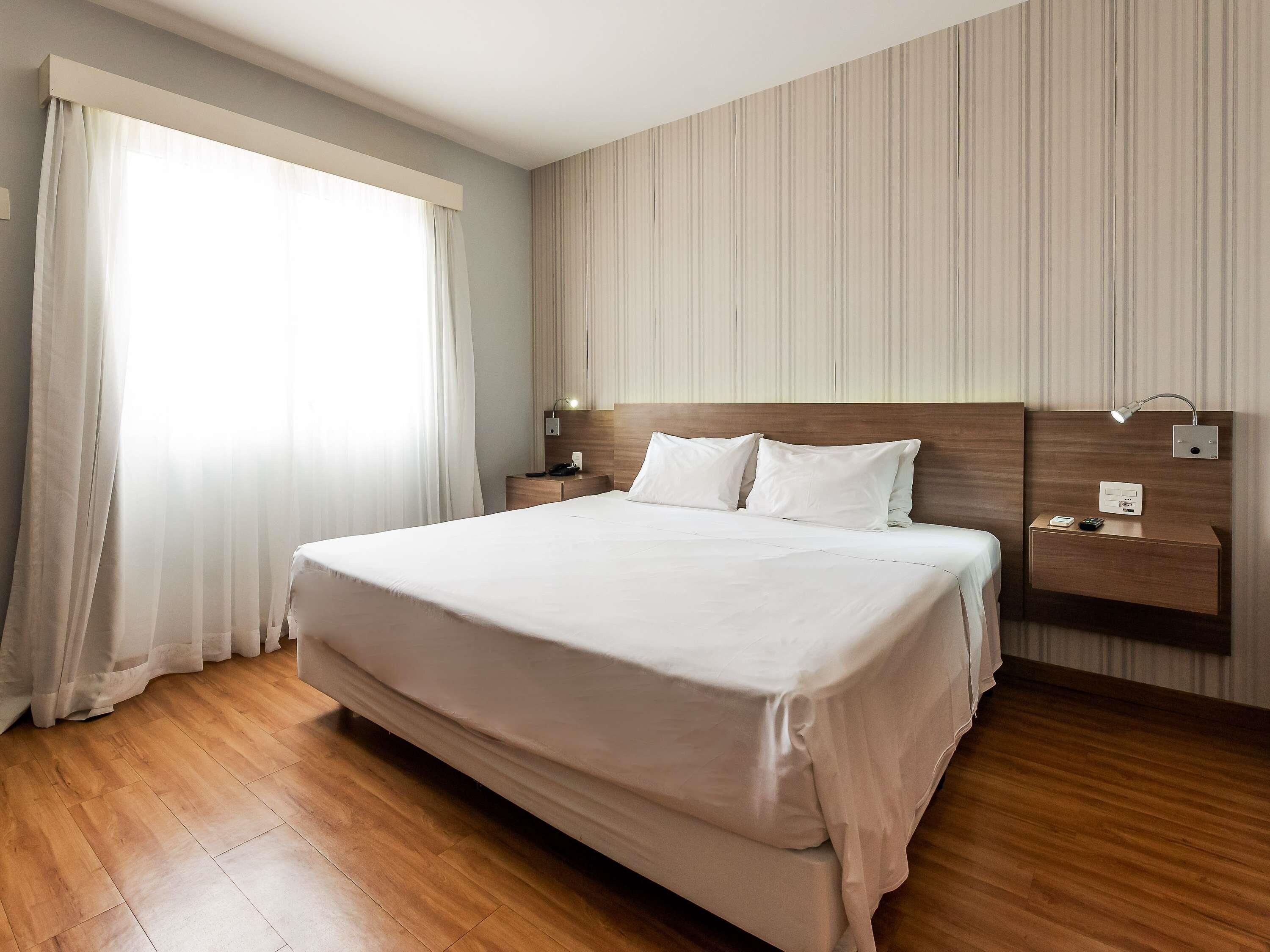 A hotel room with a large bed, wooden flooring, a nightstand with a lamp, and a wall-mounted control panel.